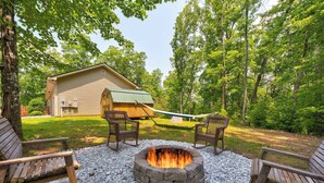 A fire pit surrounded by wooden benches in a wooded area with a building in the background.