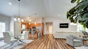 A modern living room with a large sofa, a dining area with a glass table and chairs, a kitchen with wooden cabinets, and a large plant.