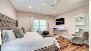 A bedroom with a large bed, a ceiling fan, a TV on the wall, and a chair.