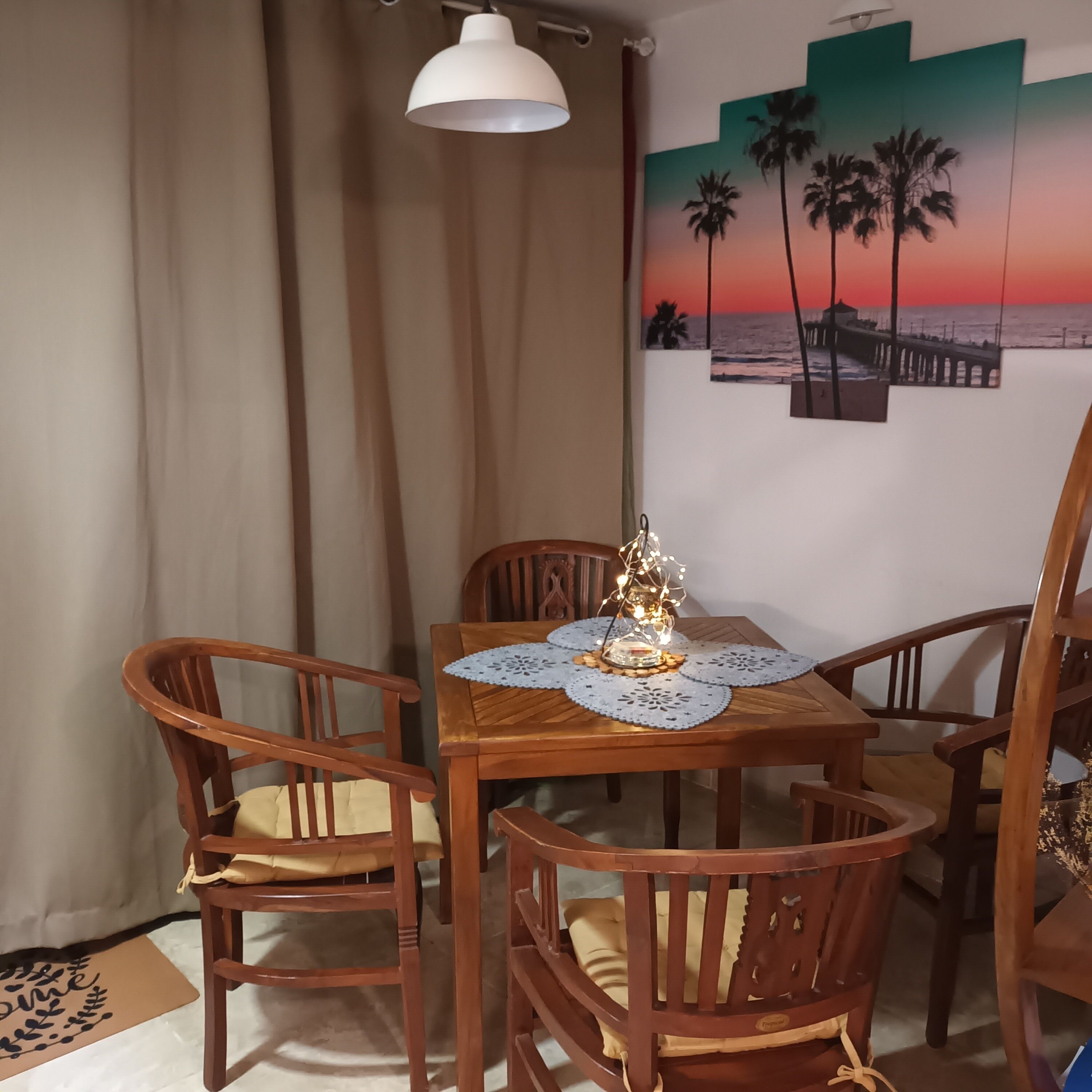 A dining area with a wooden table, four chairs, a decorative centerpiece, and a wall with a sunset painting.