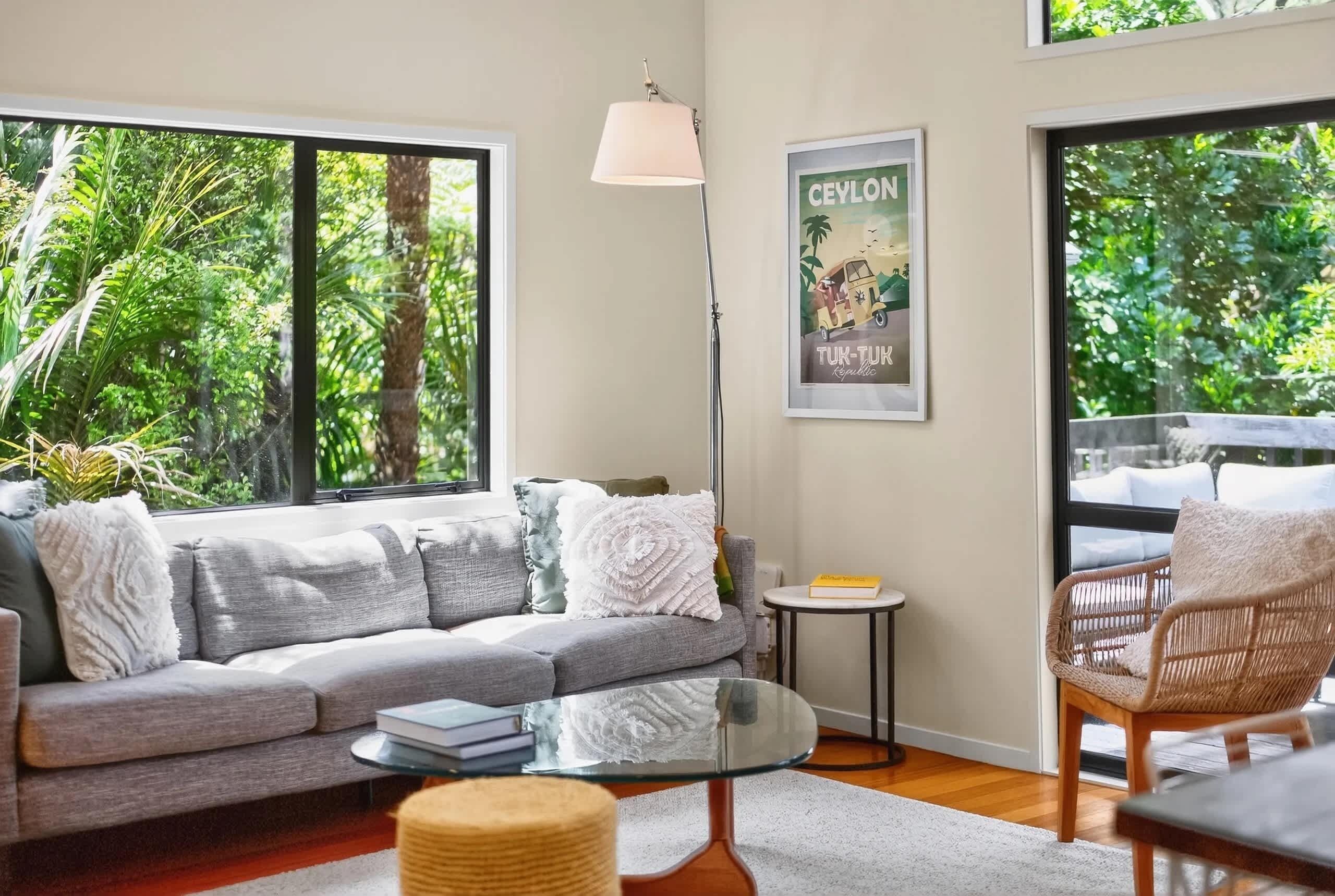 A modern living room with a grey sofa, a round glass coffee table, a wicker chair, and a floor lamp. There is a framed picture on the wall and a window with a view of greenery outside.