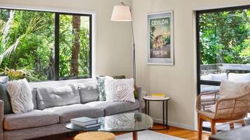 A modern living room with a grey sofa, a round glass coffee table, a wicker chair, and a floor lamp. There is a framed picture on the wall and a window with a view of greenery outside.