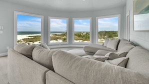 Una sala moderna con un sofá seccional grande, un ventilador de techo y ventanas amplias con vistas a la playa.
