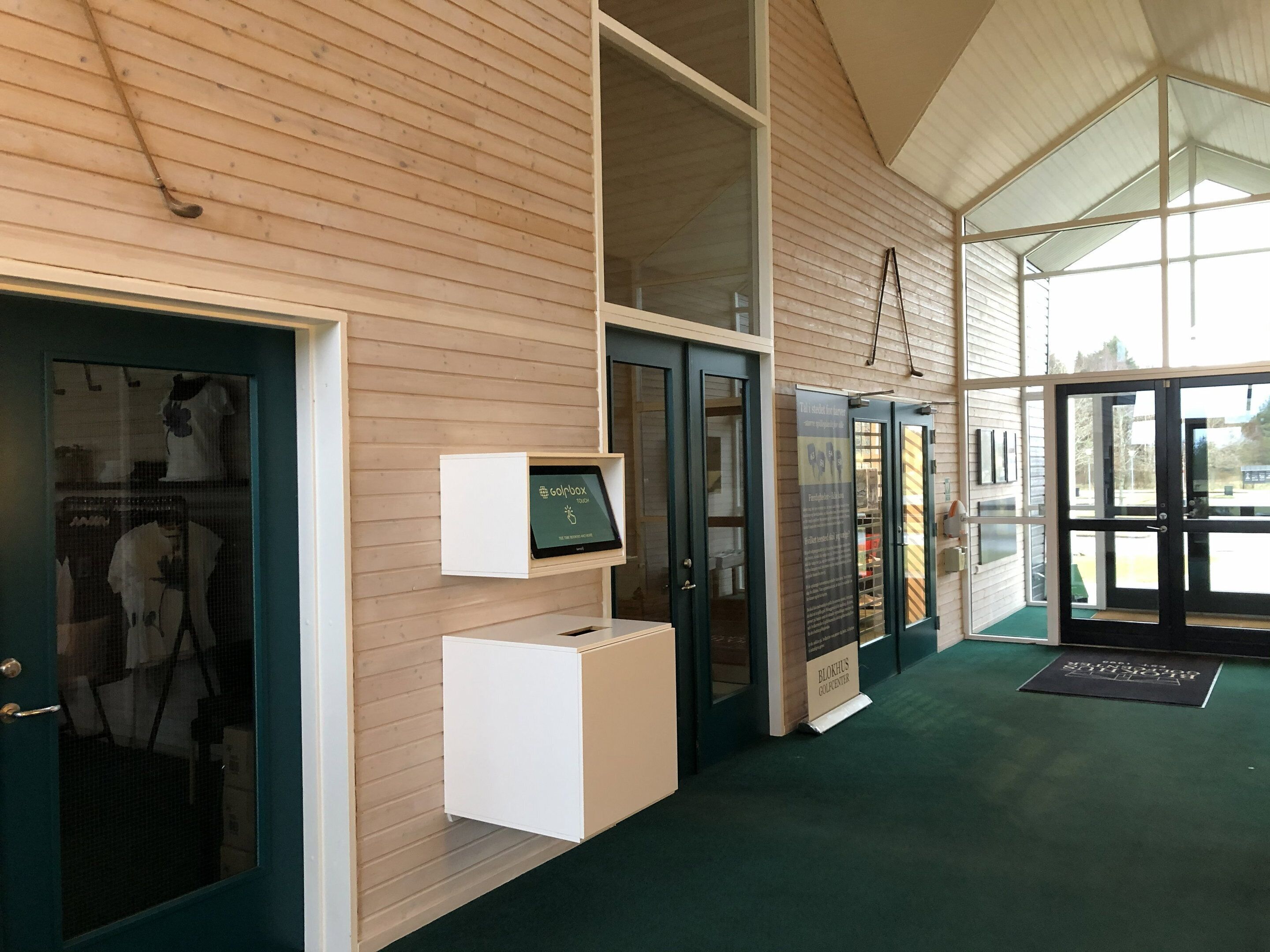 A reception area with a wooden wall, a reception desk, a digital display, and a glass door leading outside.