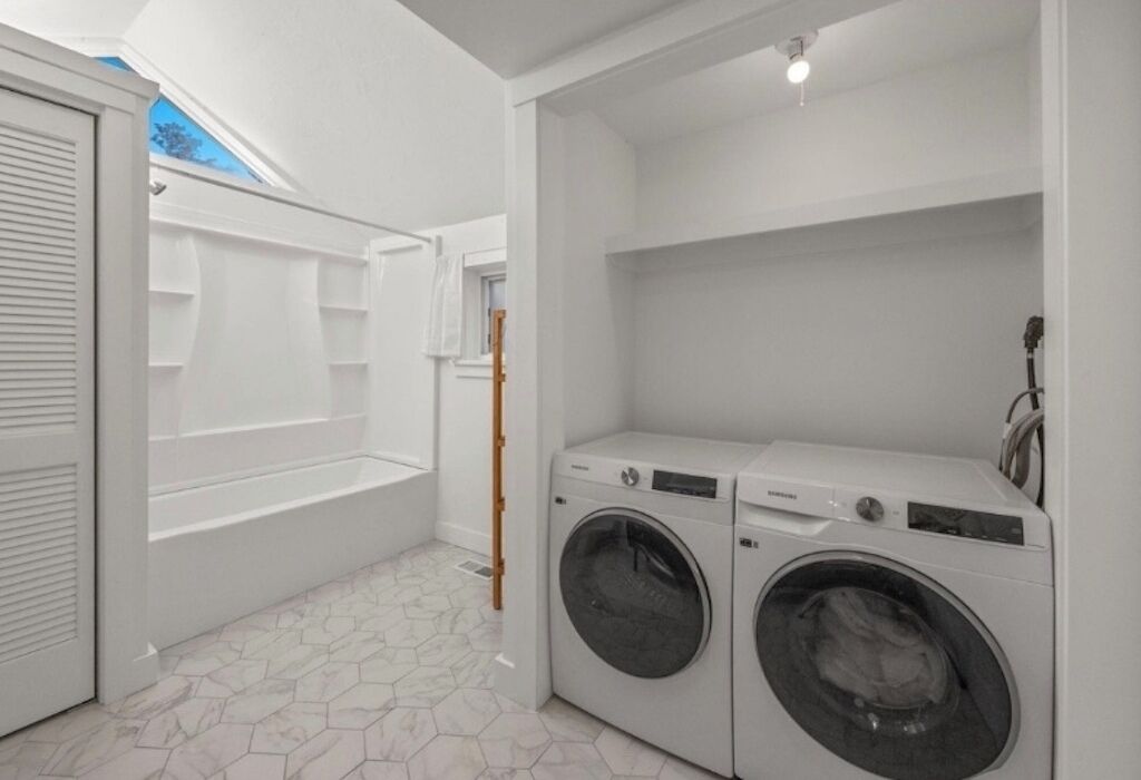 A laundry room with two front-loading washing machines, a white tiled floor, and a skylight above the shower area.