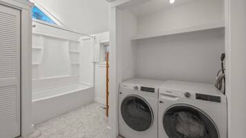 A laundry room with two front-loading washing machines, a white tiled floor, and a skylight above the shower area.