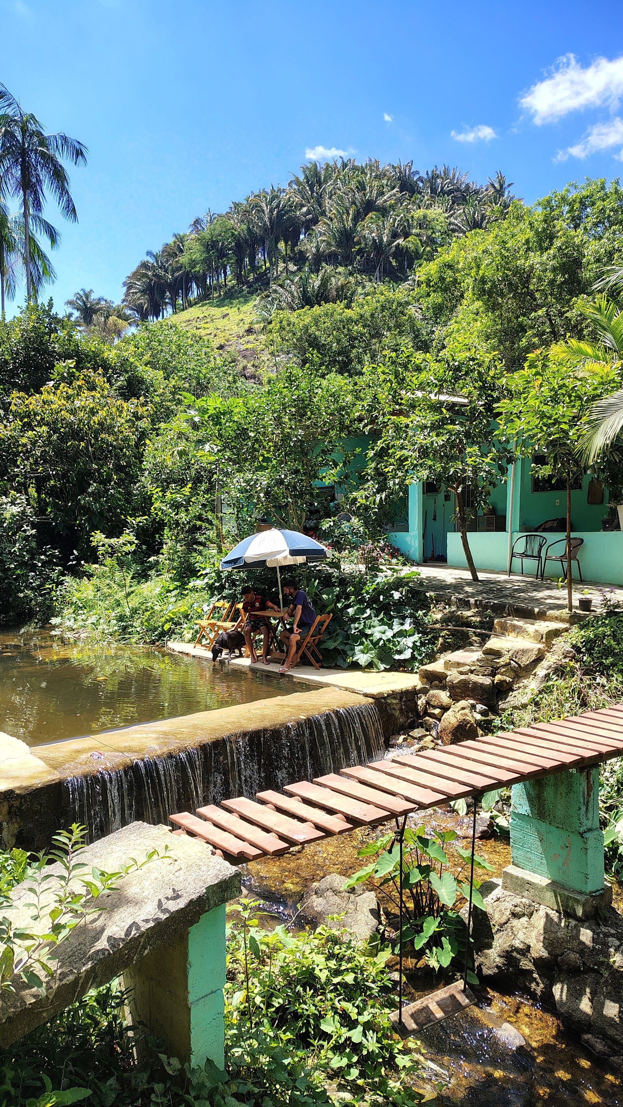 Eine ruhige Außenansicht mit einem kleinen Wasserfall, einer Holzbrücke und einer Sitzgruppe unter einem blau-weißen Sonnenschirm.