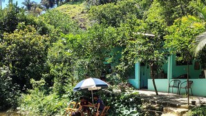 A serene outdoor setting with a small waterfall, a wooden bridge, and a seating area under a blue and white umbrella. - House in Serra do Piloto with natural pool, waterfall runs throughout the property. (Vila Muriqui)
