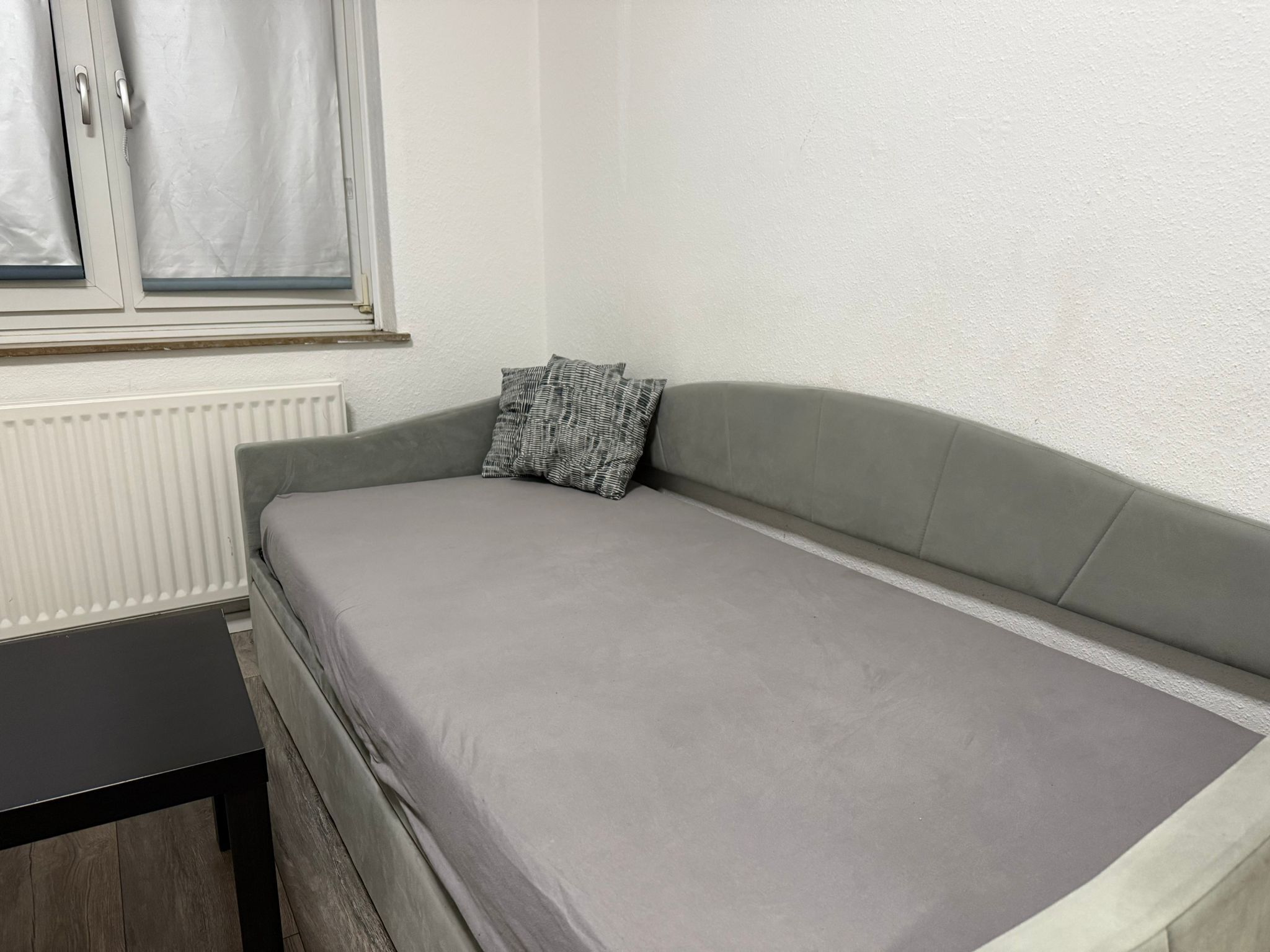 Un petit appartement d’une seule pièce, avec un canapé gris, une table noire et une fenêtre avec des stores blancs.