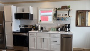 A modern kitchen with white cabinetry, stainless steel appliances, and a window with red blinds. - Cabane du quai Vacation Home (Chéticamp)