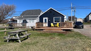 A two-story house with a porch, a picnic table, and two yellow Adirondack chairs. - Cabane du quai Vacation Home (Chéticamp)