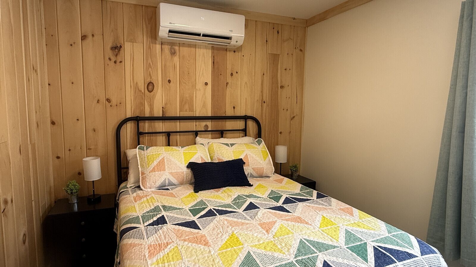 Un lit recouvert d’une couvre-lit à motifs géométriques, un oreiller foncé et deux lampes de chevet sur un mur en bois, avec un appareil de climatisation placé au-dessus.