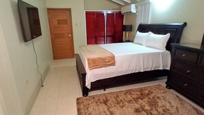 A bedroom with a bed, a dresser, a mirror, a red door, and a wall-mounted TV. - Cozy & Secure 2BR Old Harbour Retreat: All the Comforts of Your Own Home (St.Catherine)