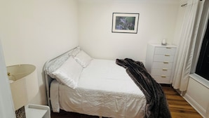 Une petite chambre à coucher, bien rangée, avec un lit simple, une table de chevet, une lampe, un radiateur et une fenêtre avec des rideaux.