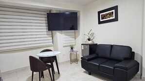 Ett vardagsrum med en svart lädersoffa, ett litet runt bord med två stolar, en platt-TV som är monterad på väggen och en inramad tavla ovanför soffan.