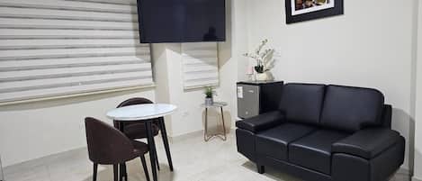 Una sala de estar con un sofá de cuero negro, una mesita redonda con dos sillas, un televisor de pantalla plana montado en la pared y un cuadro enmarcado sobre el sofá.