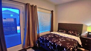 Une chambre à coucher avec un lit, des rideaux, une fenêtre avec des stores, une table de chevet et une lampe.