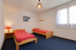 A room with two wooden beds, a small bedside table with a lamp, a framed picture on the wall, and a window with sheer curtains. - R Sidence Le Cabourg St4n (Les Deux Alpes)