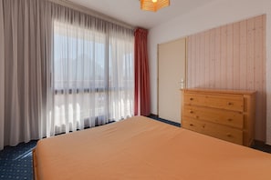 Ein Hotelzimmer mit Bett, Holzkommode, Kleiderschrank und durchsichtigen Vorhängen.