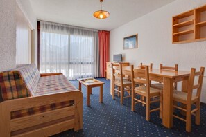 A room with a wooden sofa, a dining table with chairs, a small TV, and a window with curtains. - R Sidence Le Cabourg St4n (Les Deux Alpes)