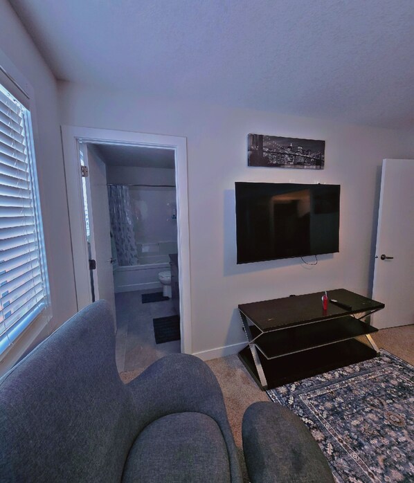 Un salon comprenant un canapé, un téléviseur fixé au mur et une porte donnant accès à une salle de bain.