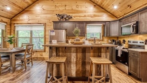 A rustic cabin interior with a wooden ceiling, a central island with stools, a kitchen with dark cabinets, and a dining area with a table and chairs. - Mountain Views, Hot Tub, Game Room & Deck (Sevierville)