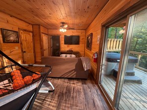 A cabin interior with a bed, a TV, a ceiling fan, and a balcony with a hot tub. - Mountain Views, Hot Tub, Game Room & Deck (Sevierville)