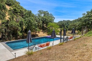 A swimming pool with lounge chairs and umbrellas, surrounded by trees and a hillside. - Lazy Coyote Ranch: 3bd 2ba, + pool on 42 acres (Sonoma County)