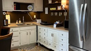 Una cucina con mobili bianchi, un frigorifero in acciaio inossidabile, un lavello e un piano di lavoro con diversi oggetti.