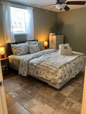 A bedroom with a bed, bedside lamps, a ceiling fan, a window with curtains, and a dresser with towels.