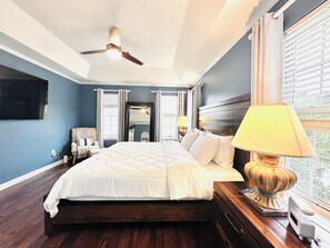 A bedroom with a bed, a ceiling fan, a nightstand with a lamp, a TV mounted on the wall, and a window with blinds. - 2027 Masters Rental House with Private Saltwater Pool & Jacuzzi bathtub (Evans)