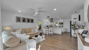 A modern living room with a beige sofa, white armchairs, and a coffee table. A kitchen with white cabinets and a dining area with white chairs is visible in the background.