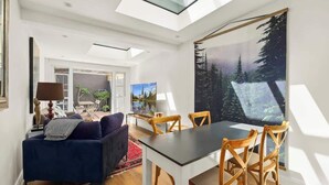 A modern living room with a dark blue sofa, wooden dining table, and chairs. A large forest-themed painting is on the wall. - Darlinghurst Sky Deck Home | City at Your Feet (Darlinghurst)