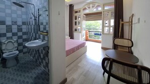 Une salle de bain avec des toilettes, un lavabo et une douche. On y trouve un lit avec une couvre-lit rose et une chaise. La pièce donne sur un balcon d’où l’on peut admirer la verdure.