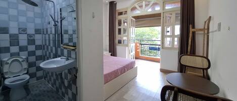 A bathroom with a toilet, sink, and shower. There is a bed with a pink bedspread and a chair. The room has a balcony with a view of greenery.
