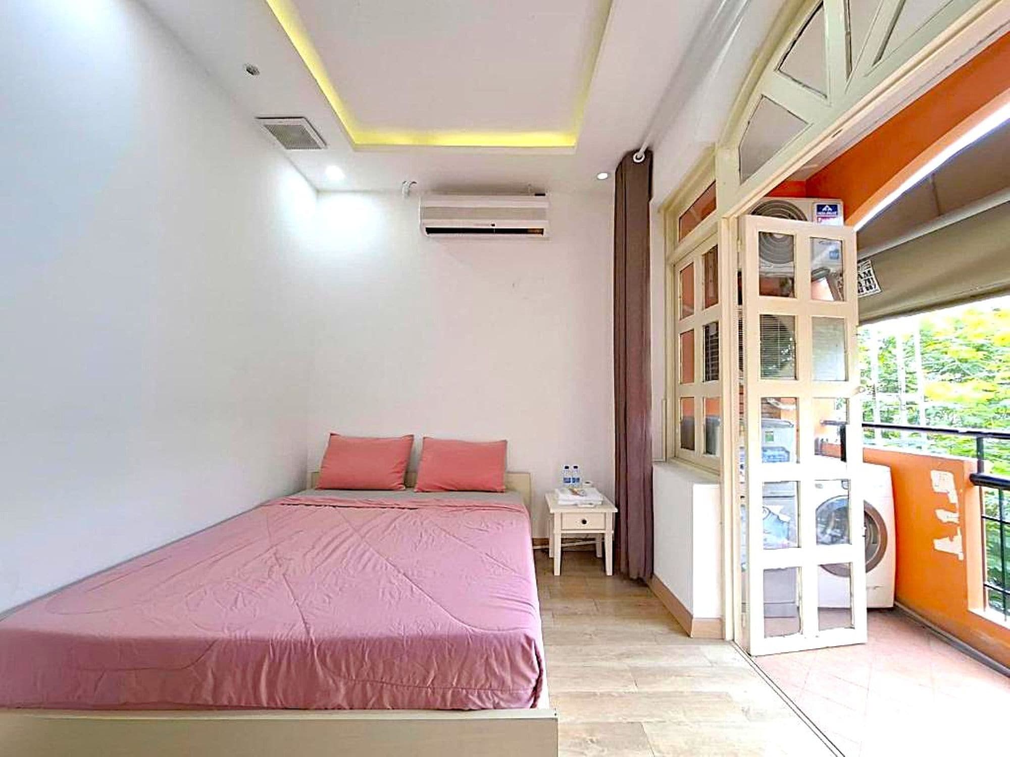 A bedroom with a bed, a nightstand, an air conditioning unit, a window with blinds, and a balcony with a washing machine.