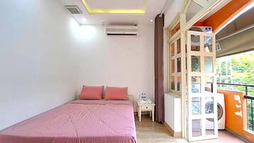 A bedroom with a bed, a nightstand, an air conditioning unit, a window with blinds, and a balcony with a washing machine.