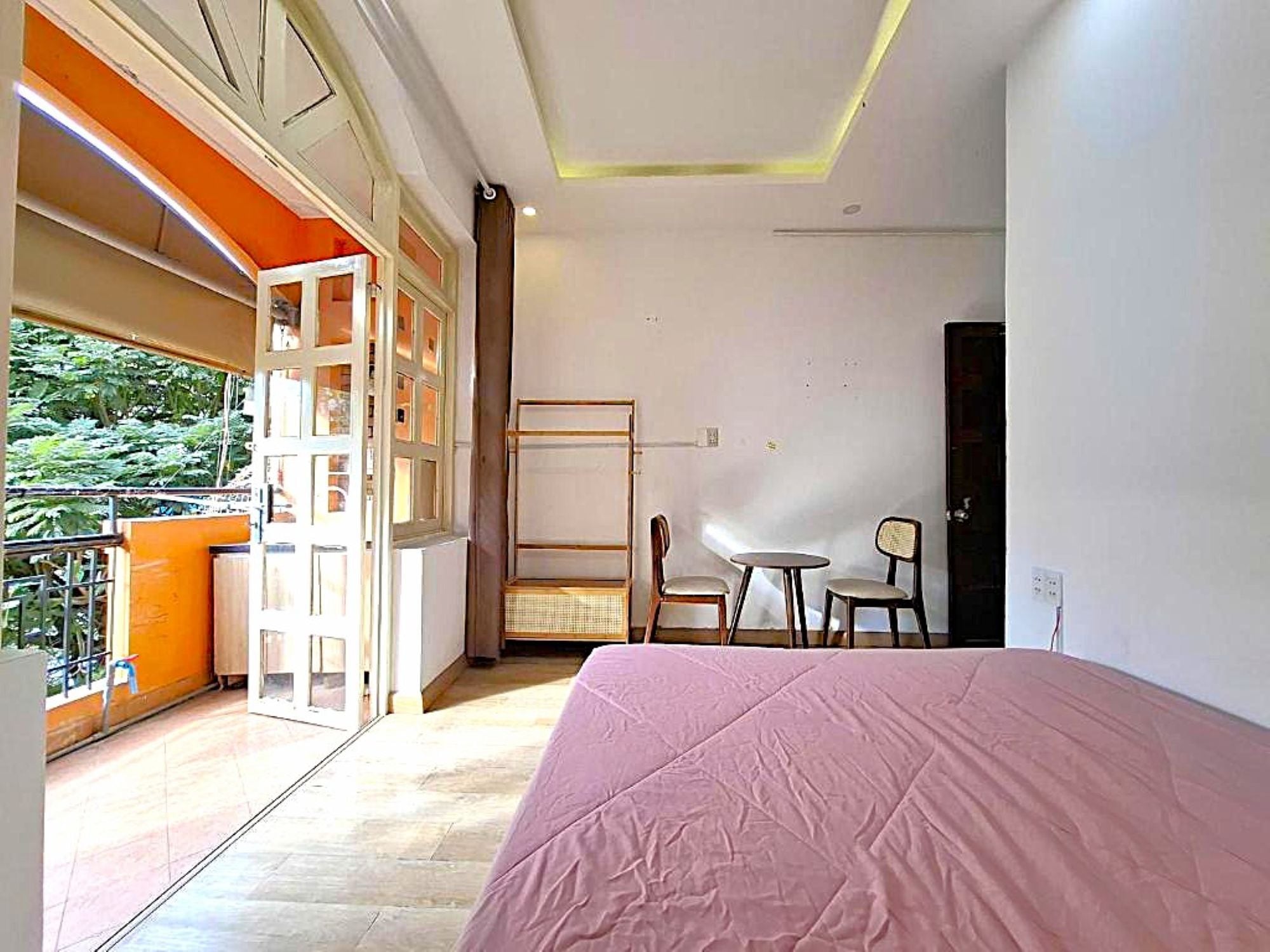 A hotel room with a bed, a small table, two chairs, a balcony with a railing, and a view of greenery.