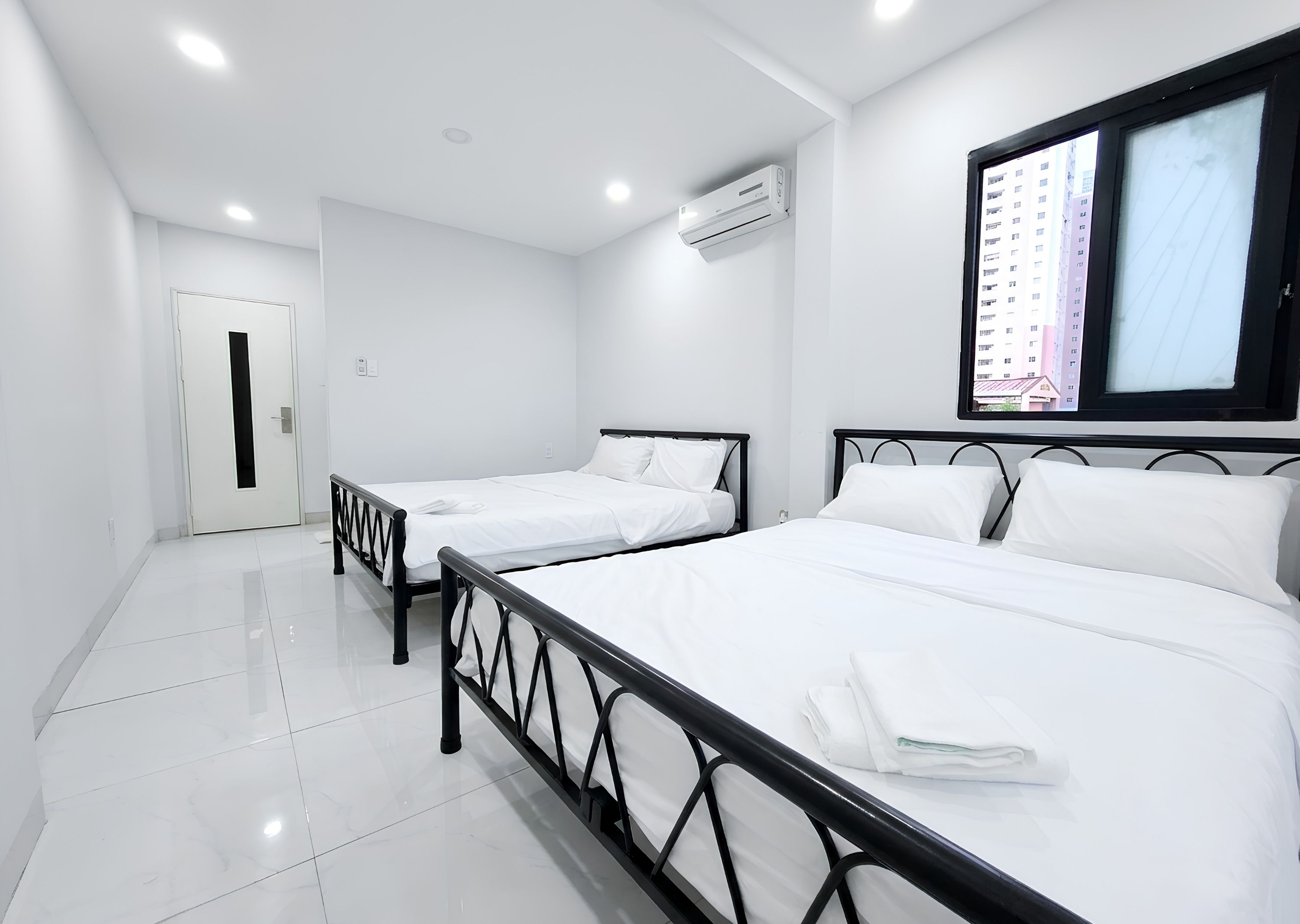 A hotel room with two single beds, a large window with a view of buildings, and a door.