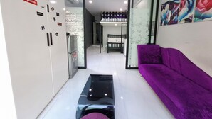 Ein modernes Wohnzimmer mit einem violetten Sofa, einem Glastisch und einem Kühlschrank mit verschiedenen Aufklebern. Im Hintergrund sieht man ein Bett mit einer weißen Matratze und einem Kopfkissen.