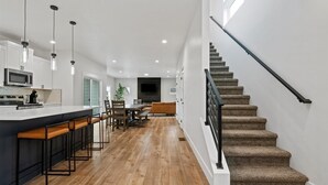Une cuisine moderne dotée d’un îlot central, de tabourets de bar et d’un coin repas avec un canapé et des chaises. Un escalier avec une rampe noire mène à l’étage supérieur.