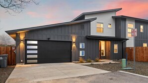 Une maison moderne à deux étages, avec une façade de couleur gris foncé, une porte de garage noire et une allée menant à une entrée fermée par un portail.