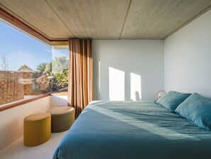 A bedroom with a large bed, a window with curtains, and a view of greenery outside. - Canaryislandshost I Famara Soul Suite (Urbanización Famara)