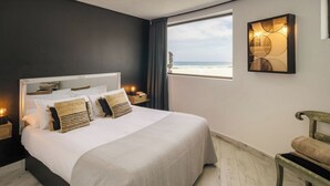 Quarto com cama grande, mesa de cabeceira com abajur, vista para a praia e luminária fixada na parede.