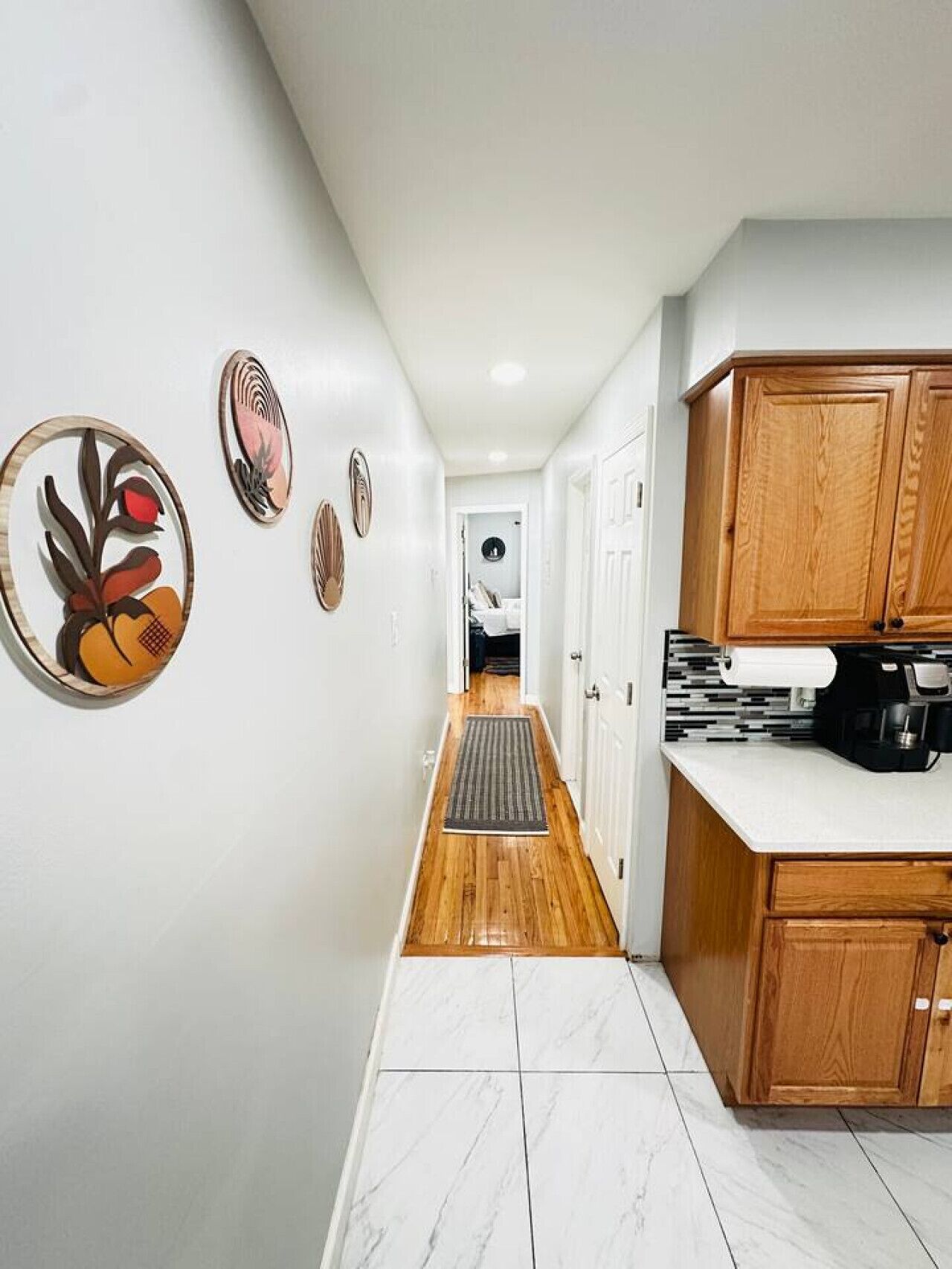 A hallway with a wooden floor, white walls, and a kitchen area with wooden cabinets and a coffee machine.