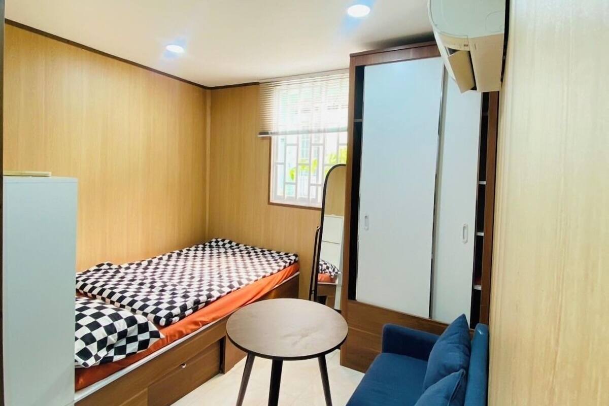 A compact room with a bed, a small table, a sofa, a wardrobe, and a window with blinds.