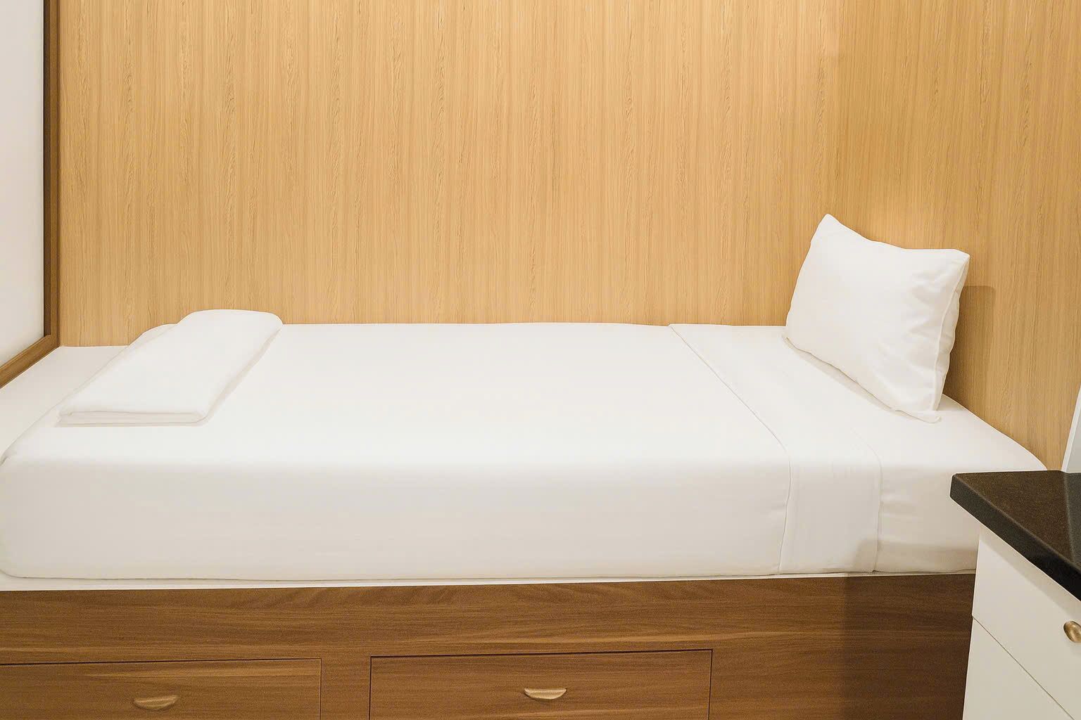 A single bed with a white mattress and pillow, wooden headboard, and a wooden nightstand with drawers.