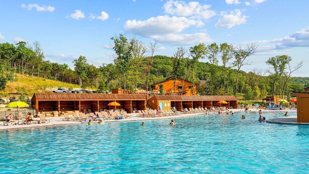 A swimming pool with lounge chairs and umbrellas, surrounded by wooden buildings and trees. - 2BR Tall Timbers Cabin | Hot Tub | Sleeps 8 | Free Tickets (Hollister)