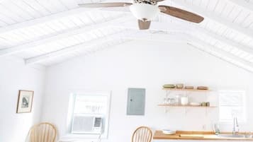 Un'ampia stanza con soffitto alto, una ventola a soffitto, un tavolo da pranzo con sedie, un angolo cottura e una finestra che lascia entrare luce naturale.