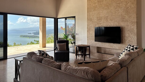 A modern living room with a large sofa, a TV mounted on a stone wall, and a view of a coastal landscape through large glass doors. - Panoramic Lake Arenal Sunset View Villa with AC (Nuevo Arenal)